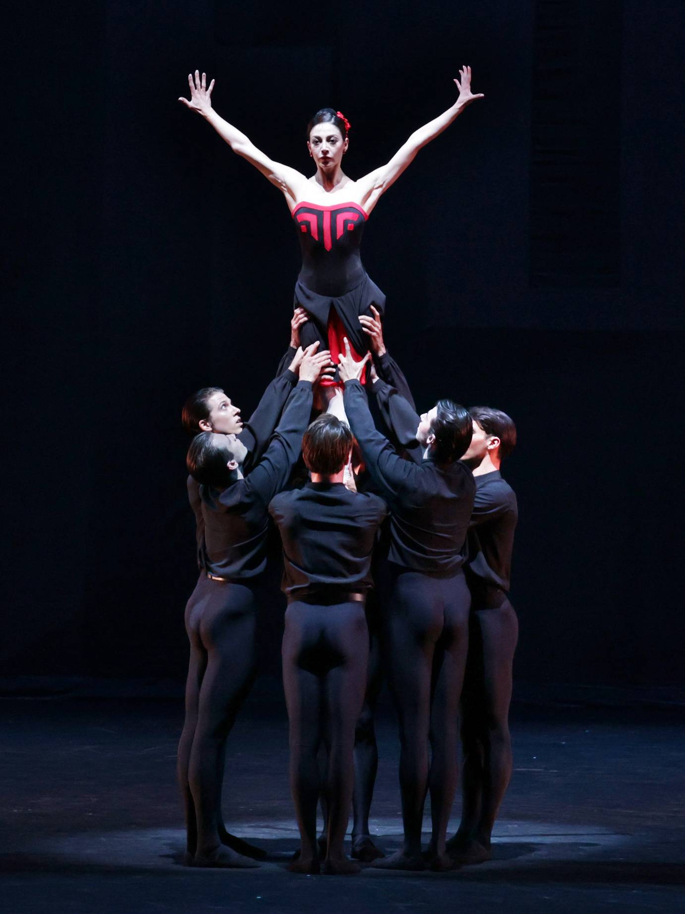 A woman costumed in black and red is held aloft by several men dressed in black.